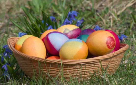 Vibrant Easter eggs in a wicker basket surrounded by grass and flowers outdoors.