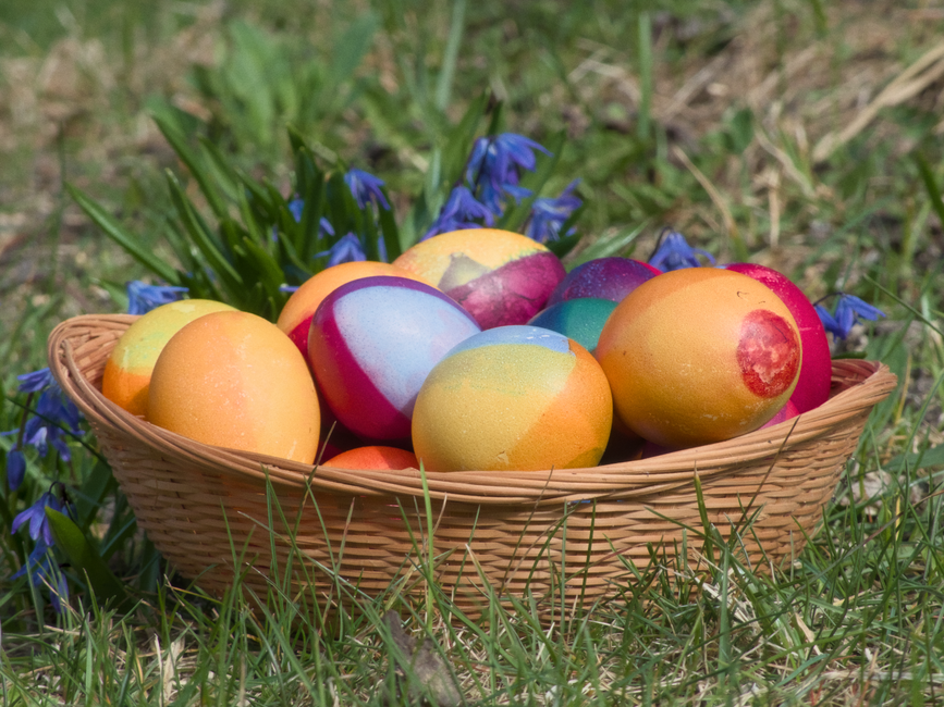 Vibrant Easter eggs in a wicker basket surrounded by grass and flowers outdoors.